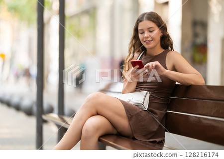 young woman sits on a bench in summer with a phone in her hands 128216174