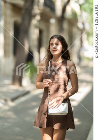 Beautiful smiling young lady wearing short dress walking shady tree lined street and relax in summer weather with sunshine outdoor 128216193