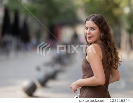 Back view of cute young girl wearing stylish short summer dress with white handbag walking along shady tree lined street enjoying spring day in Barcelona 128216244