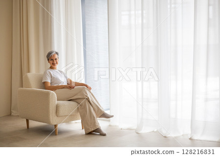 An elderly mature beautiful well-groomed woman pensioner sits near the window  128216831