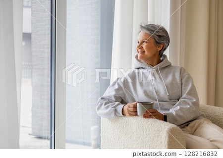 An elderly mature beautiful well-groomed woman pensioner sits near the window in a stylish chair. An elderly mature beautiful well-groomed woman pensioner sits near the window in a stylish chair. 128216832