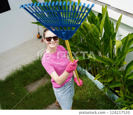 A woman raking the lawn at the backyard of her house  128217520