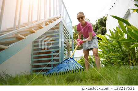 A woman raking the lawn at the backyard of her house A woman raking the lawn at the backyard of her house 128217521