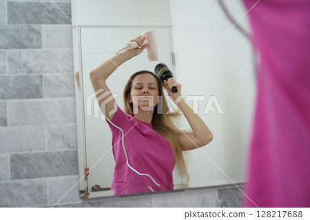 A woman is expertly styling her hair using a blow dryer and brush while in the bathroom 128217688