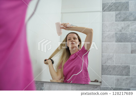A woman is expertly styling her hair using a blow dryer and brush while in the bathroom A woman is expertly styling her hair using a blow dryer and brush while in the bathroom 128217689