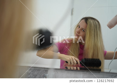 A woman is expertly styling her hair using a blow dryer and brush while in the bathroom 128217692