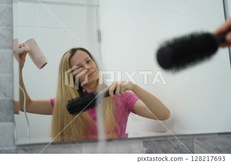 A woman is expertly styling her hair using a blow dryer and brush while in the bathroom A woman is expertly styling her hair using a blow dryer and brush while in the bathroom 128217693