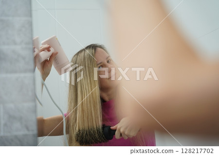 A woman is expertly styling her hair using a blow dryer and brush while in the bathroom A woman is expertly styling her hair using a blow dryer and brush while in the bathroom 128217702