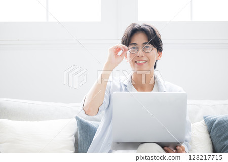 A man sitting on a sofa in front of a computer, smiling and looking at the camera 128217715