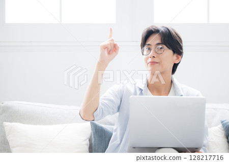 A smiling man sitting on a sofa in front of a computer looking at the camera, making a cheerful pointing pose 128217716