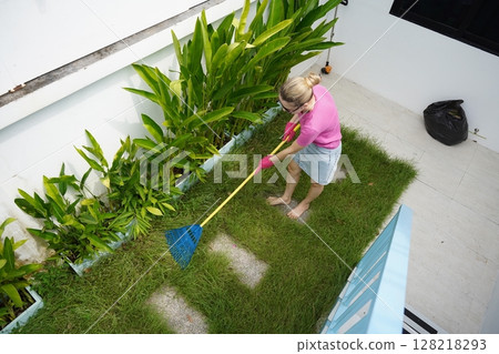 A woman raking the lawn at the backyard of her house  128218293