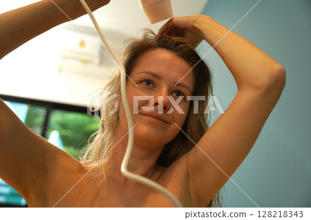 A woman is expertly styling her hair using a blow dryer and brush while in the bathroom A woman is expertly styling her hair using a blow dryer and brush while in the bathroom 128218343