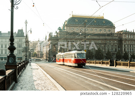 A vintage tram glides along the tracks past the National Theatre in Prague. The early morning light casts a warm glow on the historic architecture, creating a serene atmosphere. 128220965