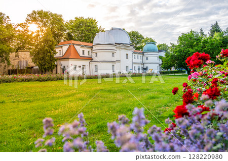 Enjoy a serene view of the Stefanik Observatory on Petrin hill in Prague as the sun sets, surrounded by lush greenery and colorful flowers in bloom. 128220980