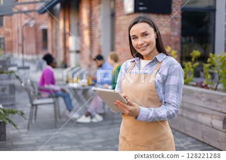 A smiling waitress in a restaurant holds a tablet, looking at the camera. Diners are in the background. 128221288