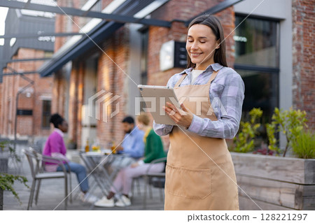 A smiling waitress takes an order at an outdoor restaurant with a tablet, overseeing customers. A smiling waitress takes an order at an outdoor restaurant with a tablet, overseeing customers. 128221297