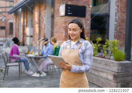 A smiling waitress takes an order on a tablet at an outdoor restaurant. A smiling waitress takes an order on a tablet at an outdoor restaurant. 128221307