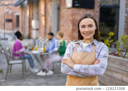 A smiling waitress in an apron poses in front of her outdoor restaurant, customers sit at tables in the background. A smiling waitress in an apron poses in front of her outdoor restaurant, customers sit at tables in the background. 128221308