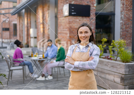 Smiling waitress standing arms crossed at a restaurant patio, with customers at a table in the background. 128221312