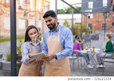 Two restaurant employees reviewing a tablet outdoors, smiling while discussing customer orders in a cafe setting. Two restaurant employees reviewing a tablet outdoors, smiling while discussing customer orders in a cafe setting. 128221314