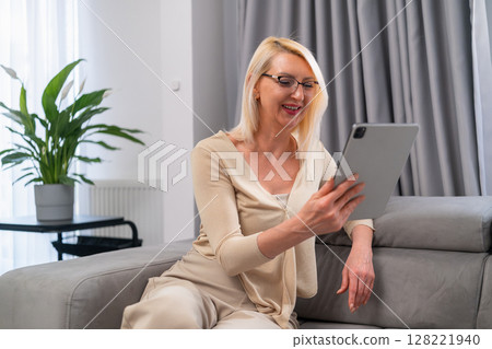 Blonde woman chatting with friend online using tablet on couch. Relaxed mature lady in casual outfit talking with her friend through video call, sitting comfortably at home. 128221940