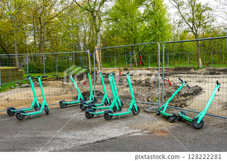 Shared Electric Scooters Parked at a Construction Site with Safety Fence and Tree Background Shared Electric Scooters Parked at a Construction Site with Safety Fence and Tree Background 128222281