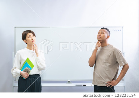 A black man and a Japanese woman thinking in front of a whiteboard in a classroom 128223154