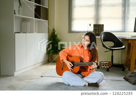 Young woman sitting on the floor, calmly playing an acoustic guitar in a cozy room Young woman sitting on the floor, calmly playing an acoustic guitar in a cozy room 128225285