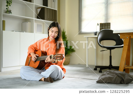 Young woman sitting on the floor, calmly playing an acoustic guitar in a cozy room 128225295
