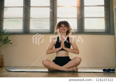 Young woman practicing yoga at home, sitting in lotus position with hands in prayer gesture 128225322