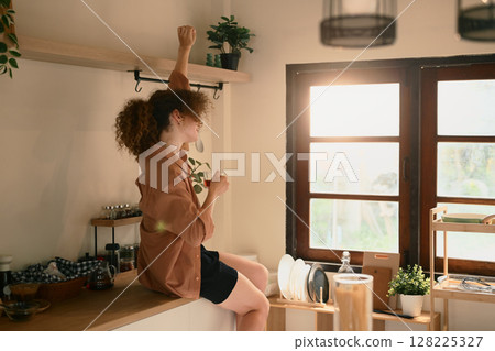 Young woman holding a cup of coffee and stretches joyfully in a sunlit kitchen 128225327