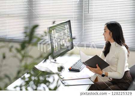 Asian woman participating in an online team meeting while reviewing documents at her desk 128225329