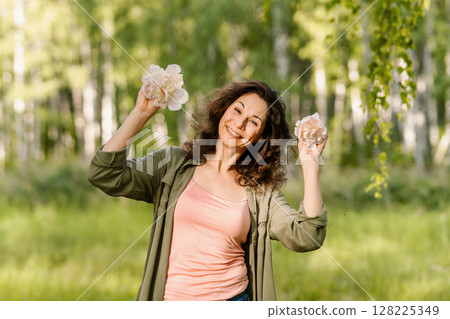 Smiling middle-aged Caucasian woman holding pink flowers near head in green summer forest. Concept of feminine energy, emotional wellness, eco lifestyle, joy and mindful connection with nature Smiling middle-aged Caucasian woman holding pink flowers near head in green summer forest. Concept of feminine energy, emotional wellness, eco lifestyle, joy and mindful connection with nature 128225349