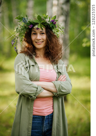 Smiling middle-aged woman with curly hair and flower wreath stands in birch forest on sunny summer day. Adult female model in casual clothes enjoys eco lifestyle, fresh air, freedom, nature connection Smiling middle-aged woman with curly hair and flower wreath stands in birch forest on sunny summer day. Adult female model in casual clothes enjoys eco lifestyle, fresh air, freedom, nature connection 128225355