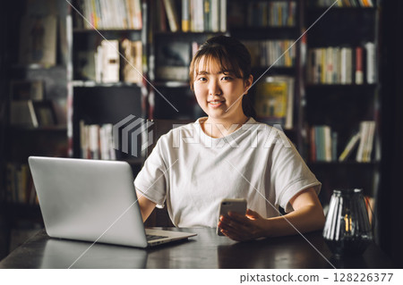 A female college student reading a book on her smartphone in a library 128226377