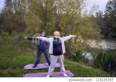 middle-aged woman teaches her old mother somatic exercises and yoga to maintain health by the river in autumn 128226393