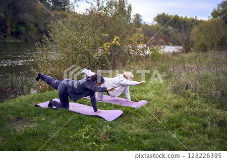 middle-aged woman teaches her old mother somatic exercises and yoga to maintain health by the river in autumn 128226395