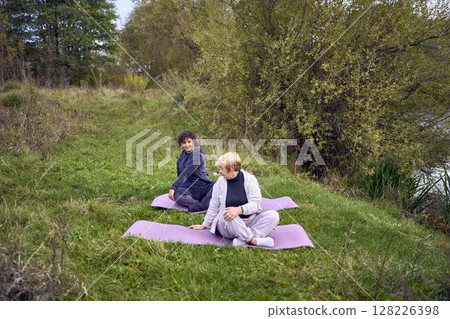 middle-aged woman teaches her old mother somatic exercises and yoga to maintain health by the river in autumn middle-aged woman teaches her old mother somatic exercises and yoga to maintain health by the river in autumn 128226398