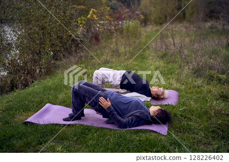 middle-aged woman teaches her old mother somatic exercises and yoga to maintain health by the river in autumn middle-aged woman teaches her old mother somatic exercises and yoga to maintain health by the river in autumn 128226402