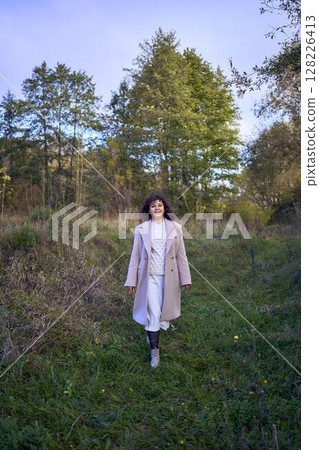 middle-aged woman with dark curly hair in light beige suit and coat enjoying nature in autumn forest middle-aged woman with dark curly hair in light beige suit and coat enjoying nature in autumn forest 128226413