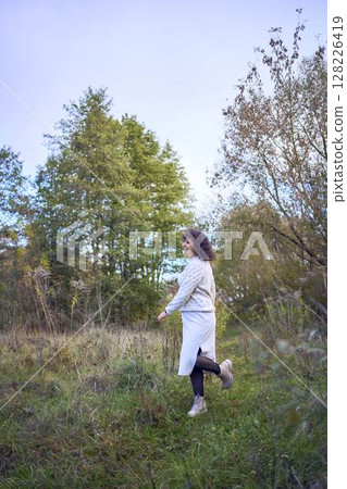 middle-aged woman with dark curly hair in light beige suit enjoying nature in autumn forest middle-aged woman with dark curly hair in light beige suit enjoying nature in autumn forest 128226419