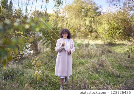 middle-aged woman with dark curly hair in light beige suit and coat enjoying nature in autumn forest middle-aged woman with dark curly hair in light beige suit and coat enjoying nature in autumn forest 128226426
