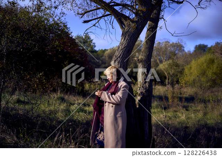 elderly woman with gray hair in light beige coat enjoying nature in autumn forest 128226438