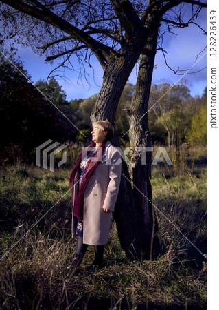 elderly woman with gray hair in light beige coat enjoying nature in autumn forest 128226439