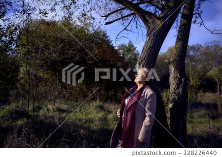 elderly woman with gray hair in light beige coat enjoying nature in autumn forest 128226440