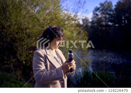 middle-aged woman in wireless headphones drinking coffee, tea from a thermos on a walk in the autumn forest middle-aged woman in wireless headphones drinking coffee, tea from a thermos on a walk in the autumn forest 128226470