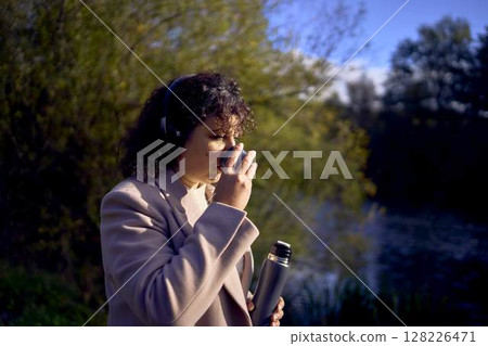 middle-aged woman in wireless headphones drinking coffee, tea from a thermos on a walk in the autumn forest 128226471