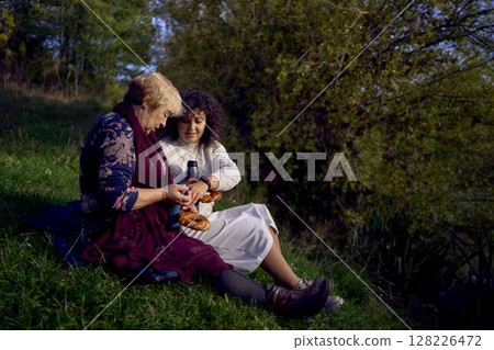 middle-aged daughter and old mother eating pastries and drinking tea from thermos cups on a picnic in the autumn forest 128226472