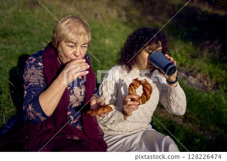 middle-aged daughter and old mother eating pastries and drinking tea from thermos cups on a picnic in the autumn forest middle-aged daughter and old mother eating pastries and drinking tea from thermos cups on a picnic in the autumn forest 128226474