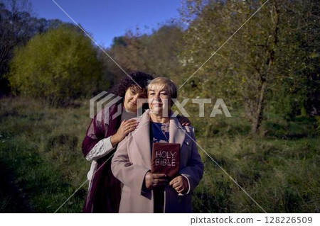 middle-aged woman and elderly mother with holy bible in hands praying for peace in autumn forest 128226509